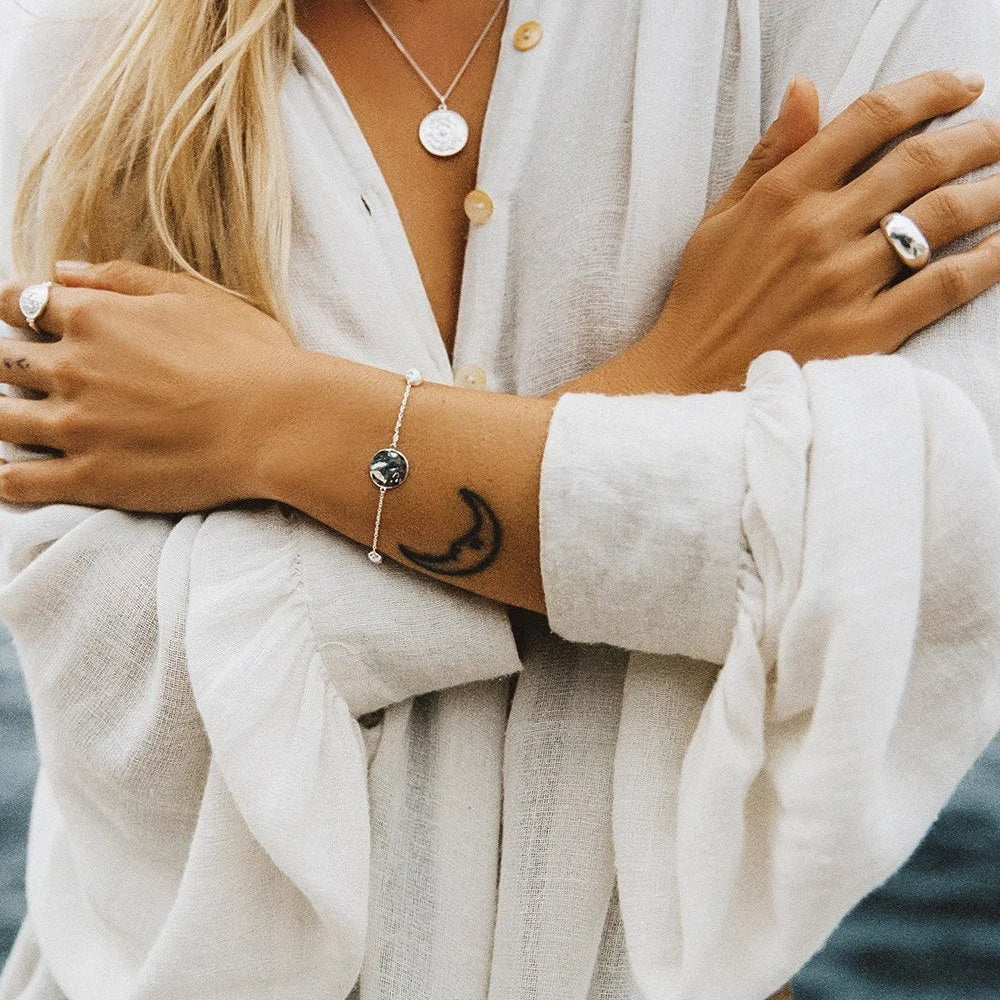 A close-up lifestyle shot of a woman wearing a delicate silver chain bracelet with a small crescent moon charm, styled with a white linen button-up shirt.