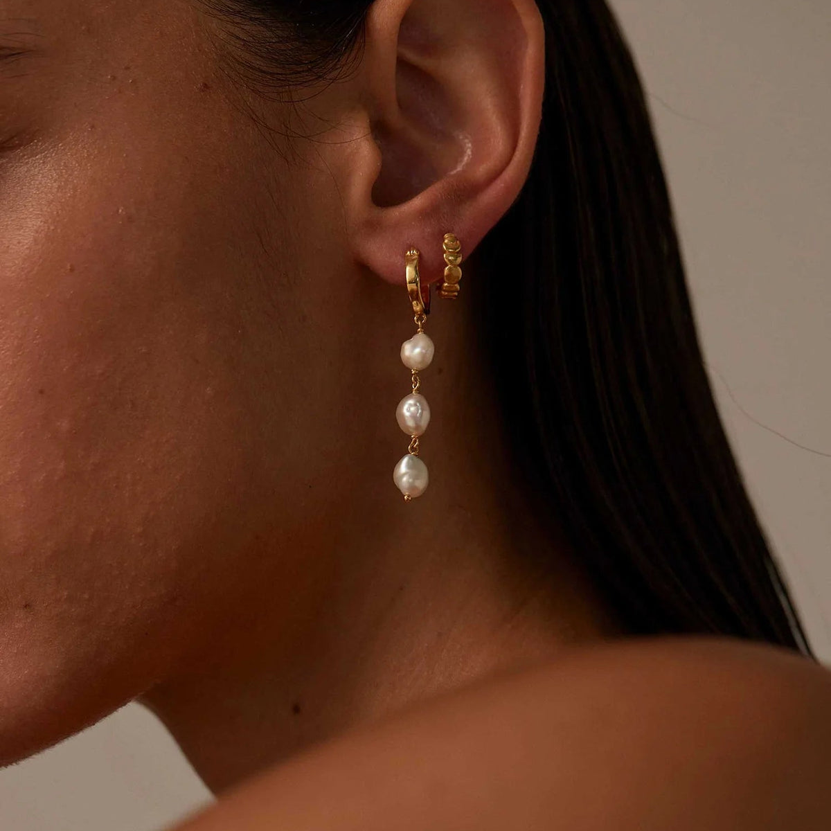 A close-up of a model's ear wearing a gold huggie hoop earring with three dangling freshwater pearls, set against a warm neutral background.