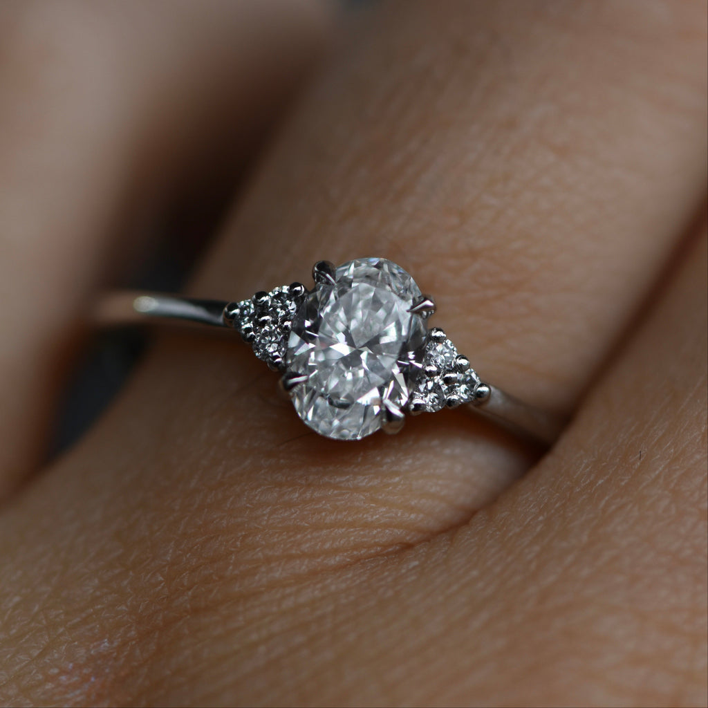 A close-up of a platinum engagement ring with a oval cut center stone and small side stones, worn on a finger.