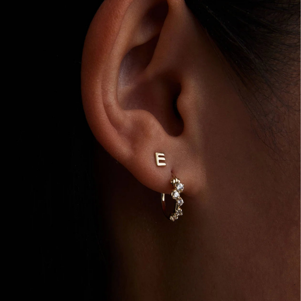 Close-up of a person's ear wearing a small gold hoop earring with clear stones and a single gold initial 'E' stud earring, against a dark background.