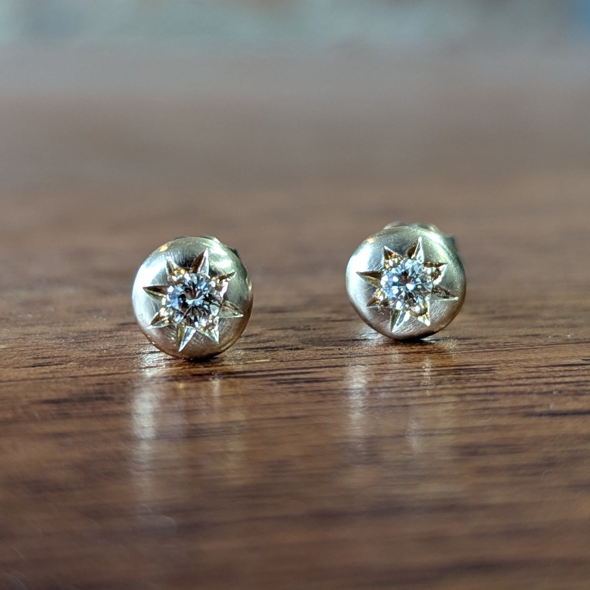 A pair of round gold stud earrings with diamonds, resting on a dark wooden surface.
