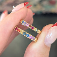 Gold earrings with colorful gemstones held between fingers against a blurred background