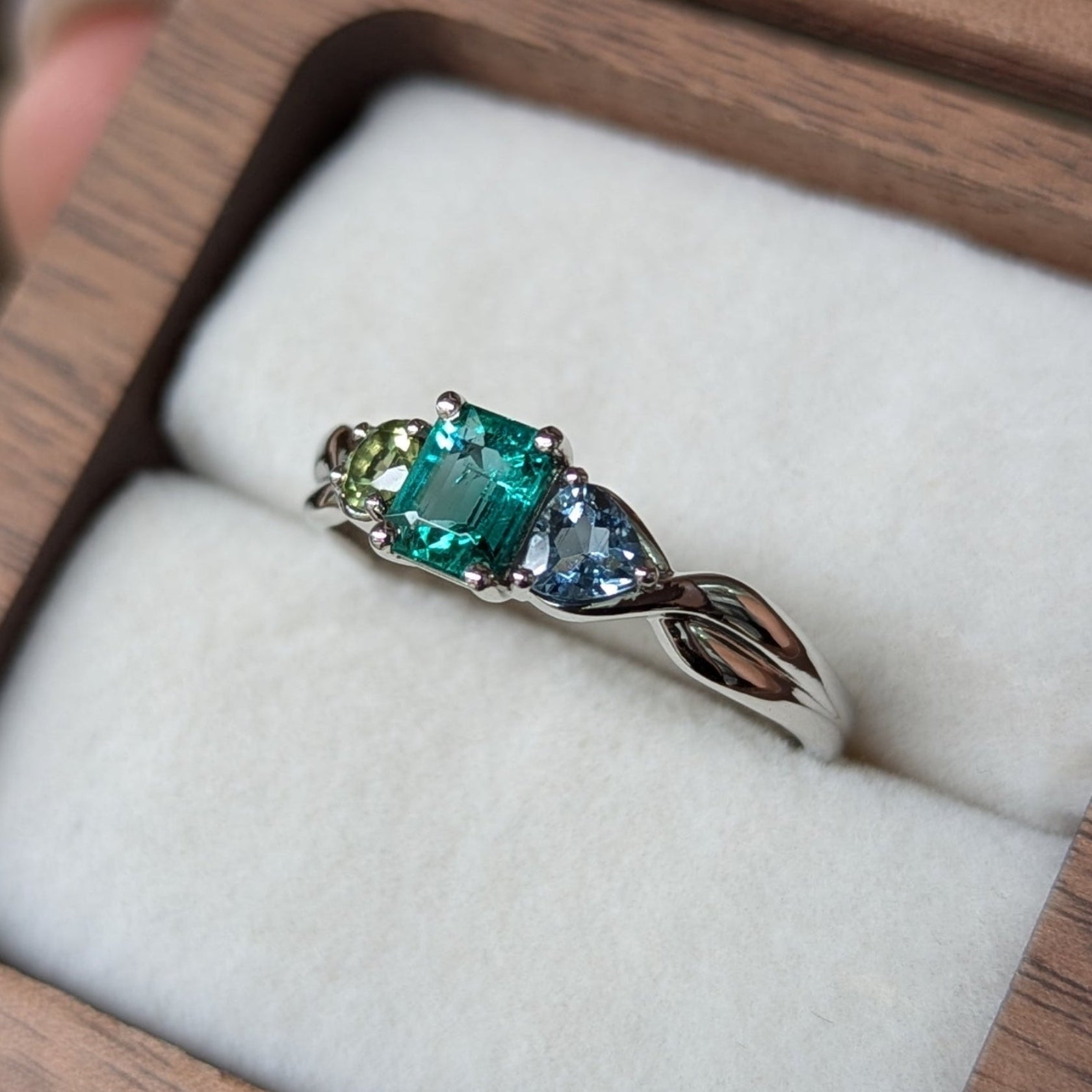 Trilogy style ring with Round Peridot, Emerald Cut Emerald and Trillion Cut Aquamarine gemstone in a wooden box