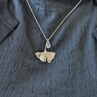 Person wearing a dark jacket with a necklace featuring a gingko leaf and moonstone pendant.