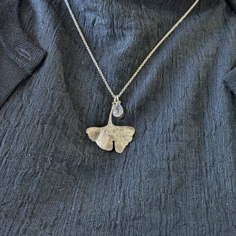 Person wearing a dark jacket with a necklace featuring a gingko leaf and moonstone pendant.