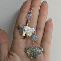 Hand holding a pair of silver earrings with a light-colored moonstone against a plain background