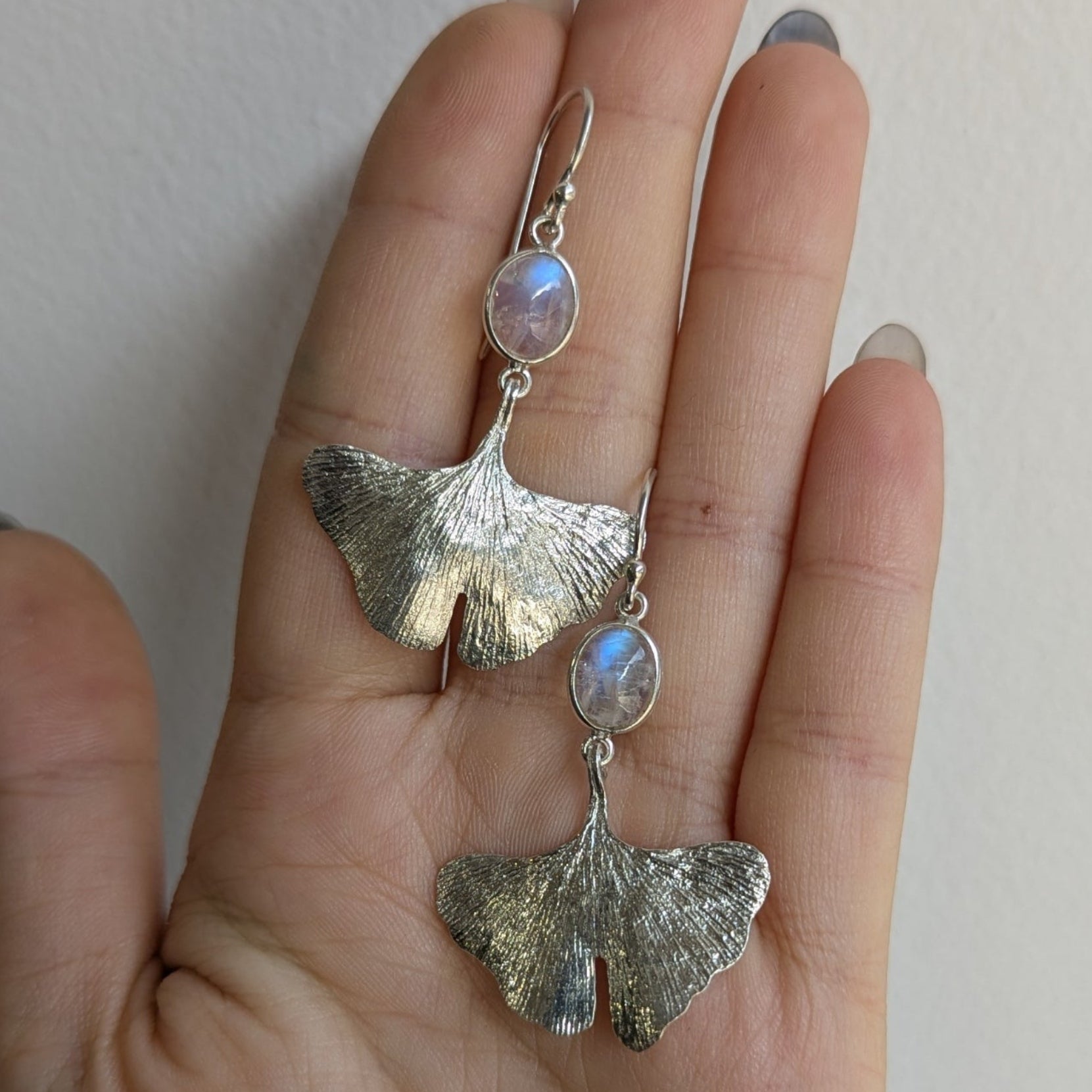 Hand holding a pair of silver earrings with a light-colored moonstone against a plain background