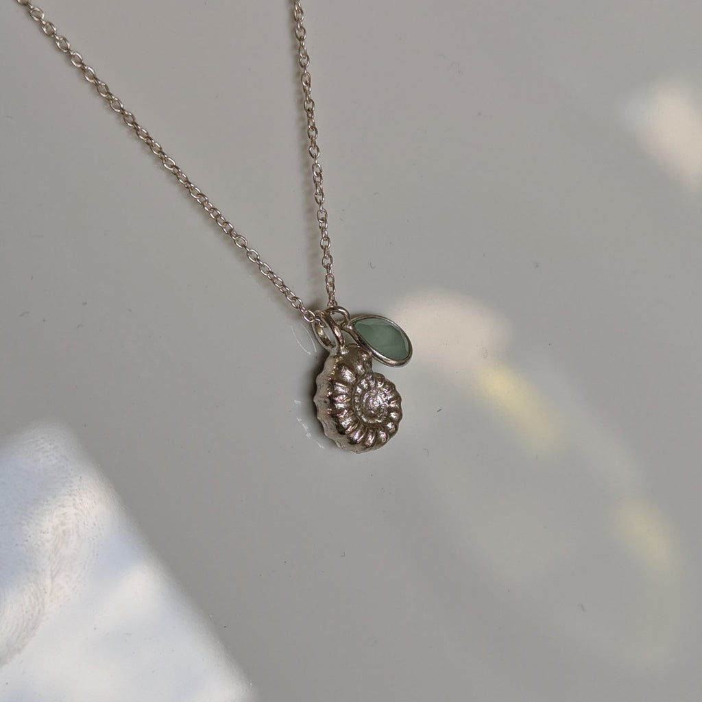 A delicate silver chain necklace with a small spiral shell pendant and a tiny green stone accent, resting on a light gray surface.