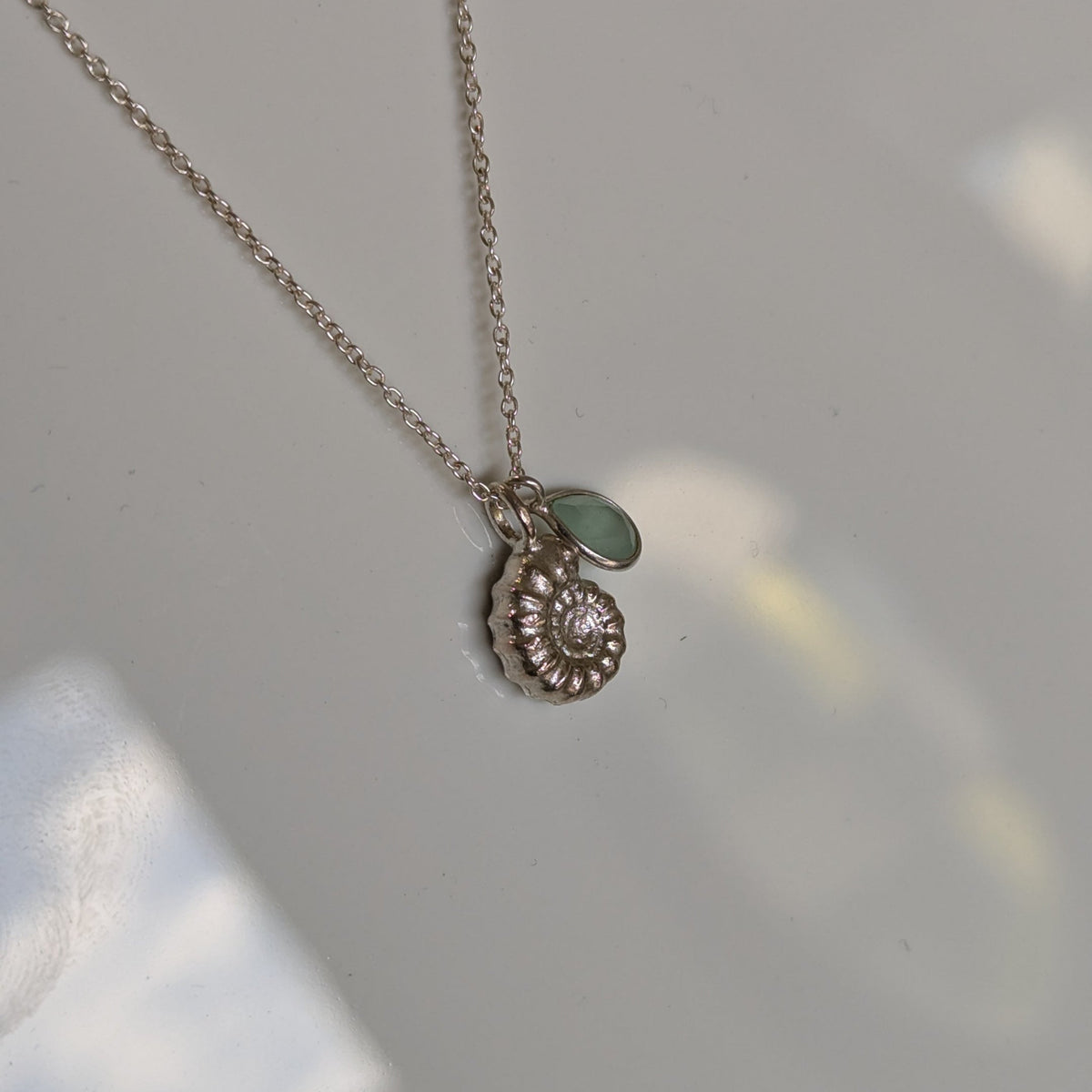 A delicate silver chain necklace with a small spiral shell pendant and a tiny green stone accent, resting on a light gray surface.