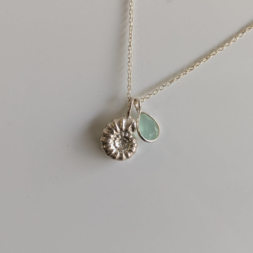 A delicate silver necklace featuring a textured, flower-like round pendant and a small, teardrop-shaped light green agate pendant, displayed on a plain light gray background.