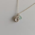 A delicate silver necklace featuring a textured, flower-like round pendant and a small, teardrop-shaped light green agate pendant, displayed on a plain light gray background.