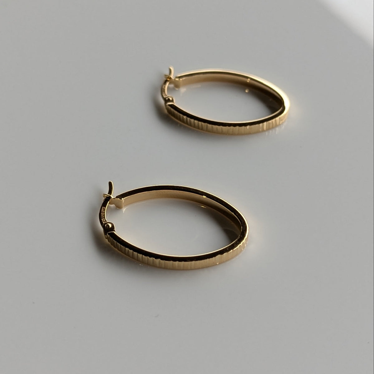 A pair of textured gold hoop earrings resting on a clean, light surface with a soft diagonal shadow.