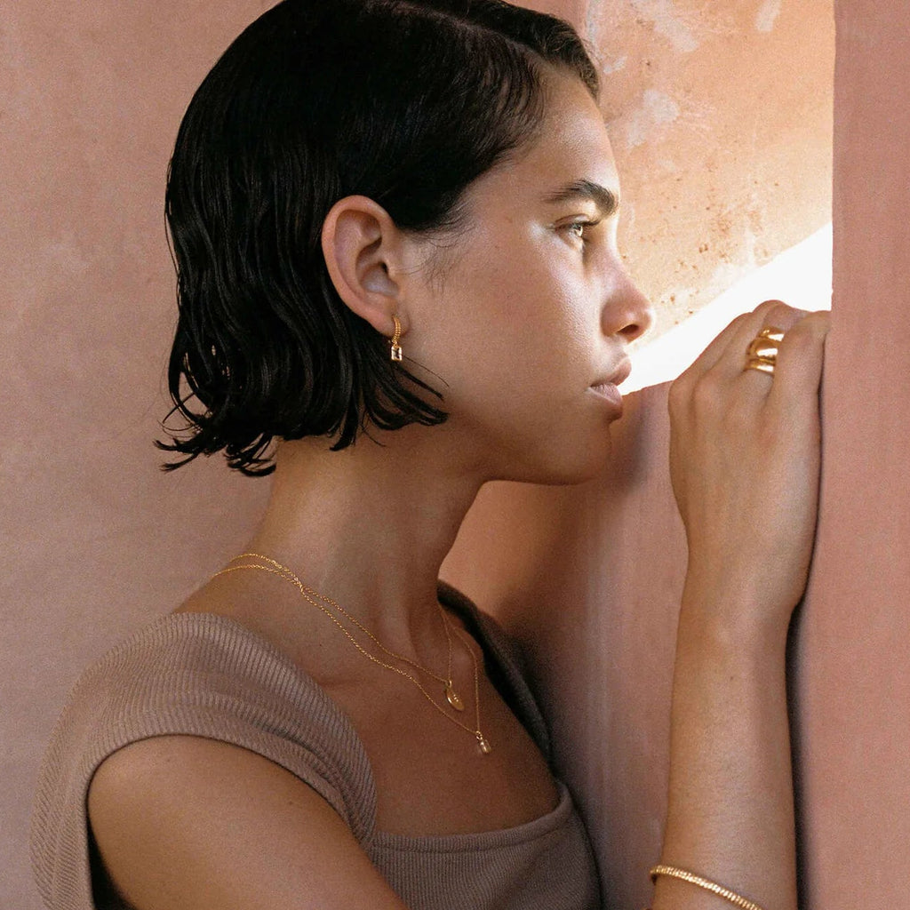 A woman with short dark hair leans against a textured pink wall, wearing delicate gold jewelry including a small gold hoop earring, a thin gold necklace, and stacked gold bangles.